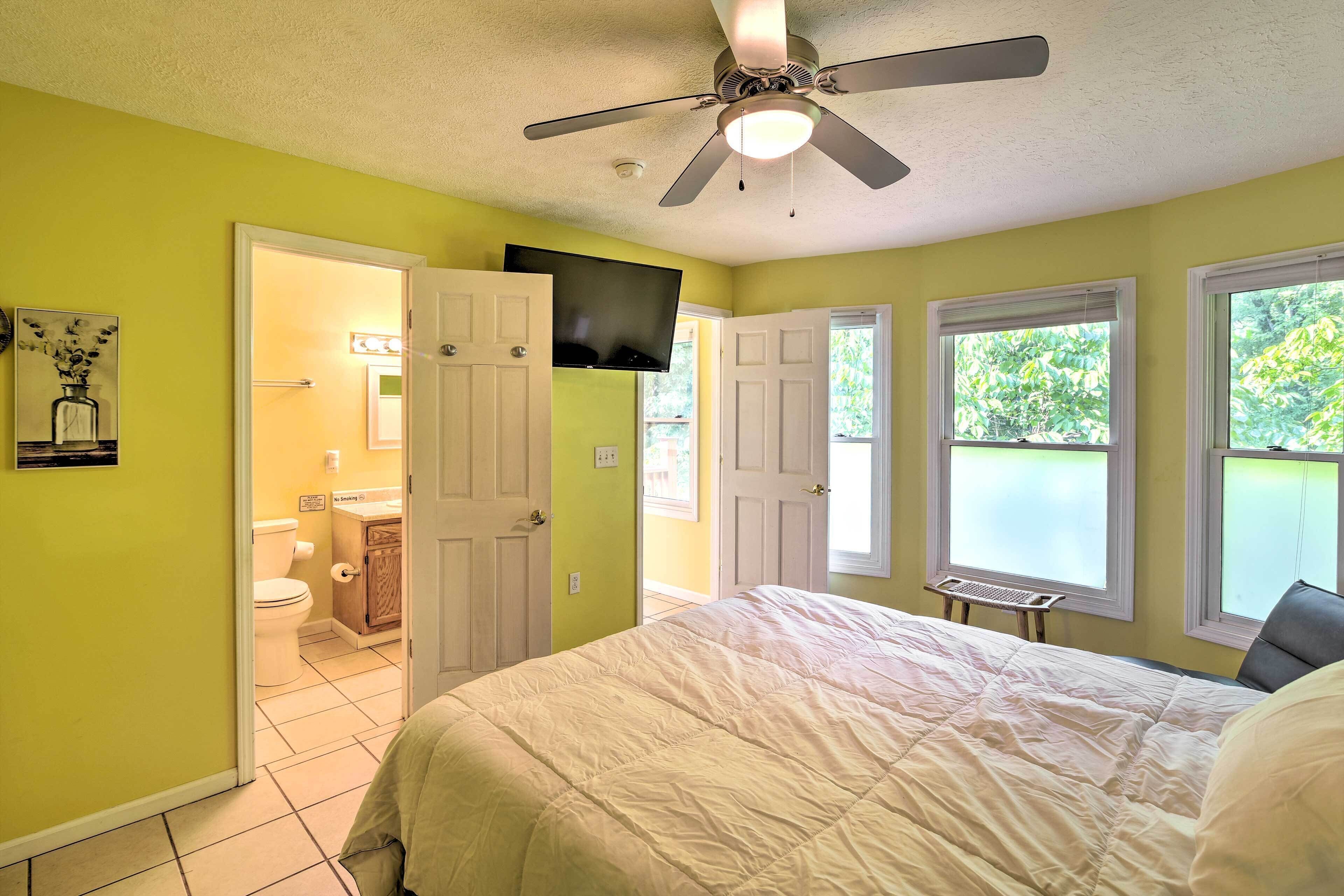 A bedroom with a bed, a ceiling fan, a television, and a window with a view of greenery.