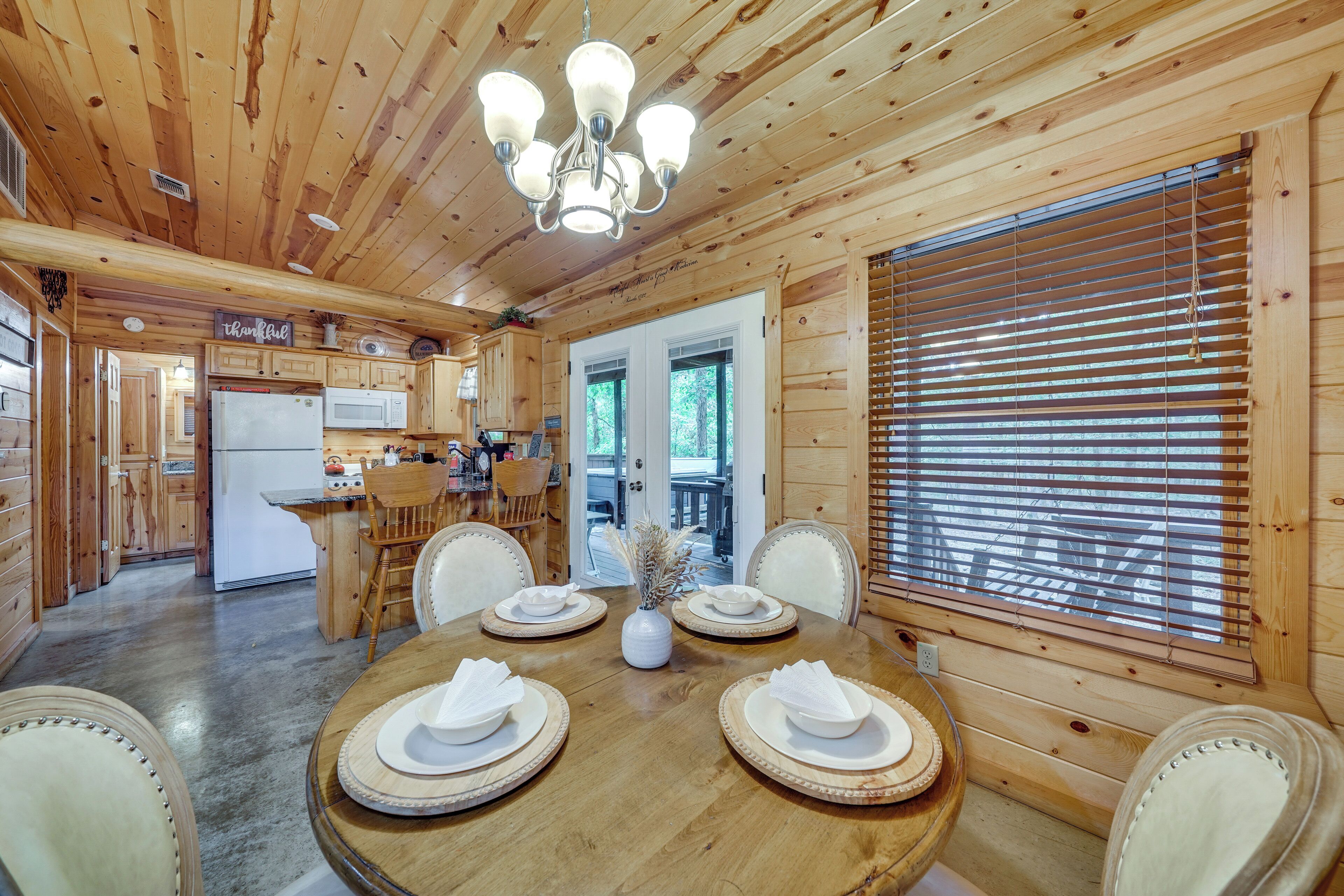 Interior de una cabaña de madera con una mesa de comedor para cuatro personas, una zona de cocina y un gran ventanal con persianas.