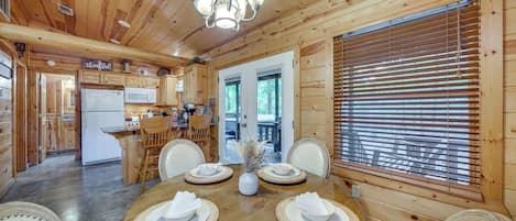 A wooden cabin interior with a dining table set for four, a kitchen area, and a large window with blinds.