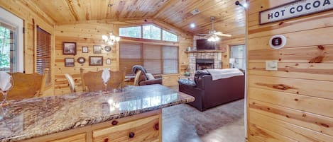 A wooden cabin interior with a kitchen area, a dining table, and a living room with a fireplace.