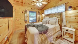 Une chambre à coucher dans un chalet en bois, comprenant un lit, une table de chevet, un ventilateur de plafond, une fenêtre avec des stores et un miroir.