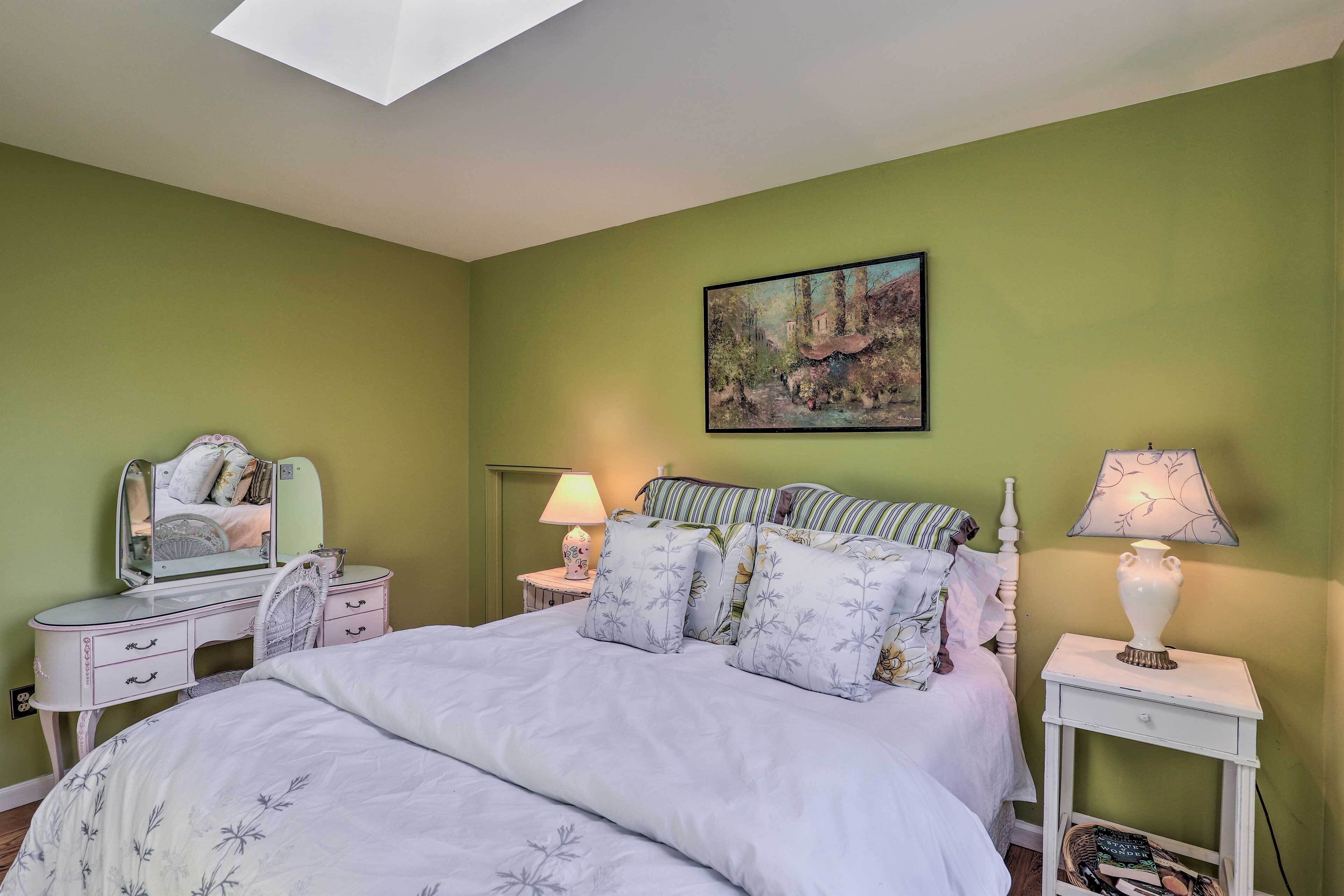A bedroom with a bed, a vanity with a mirror, two bedside tables with lamps, and a painting on the wall.
