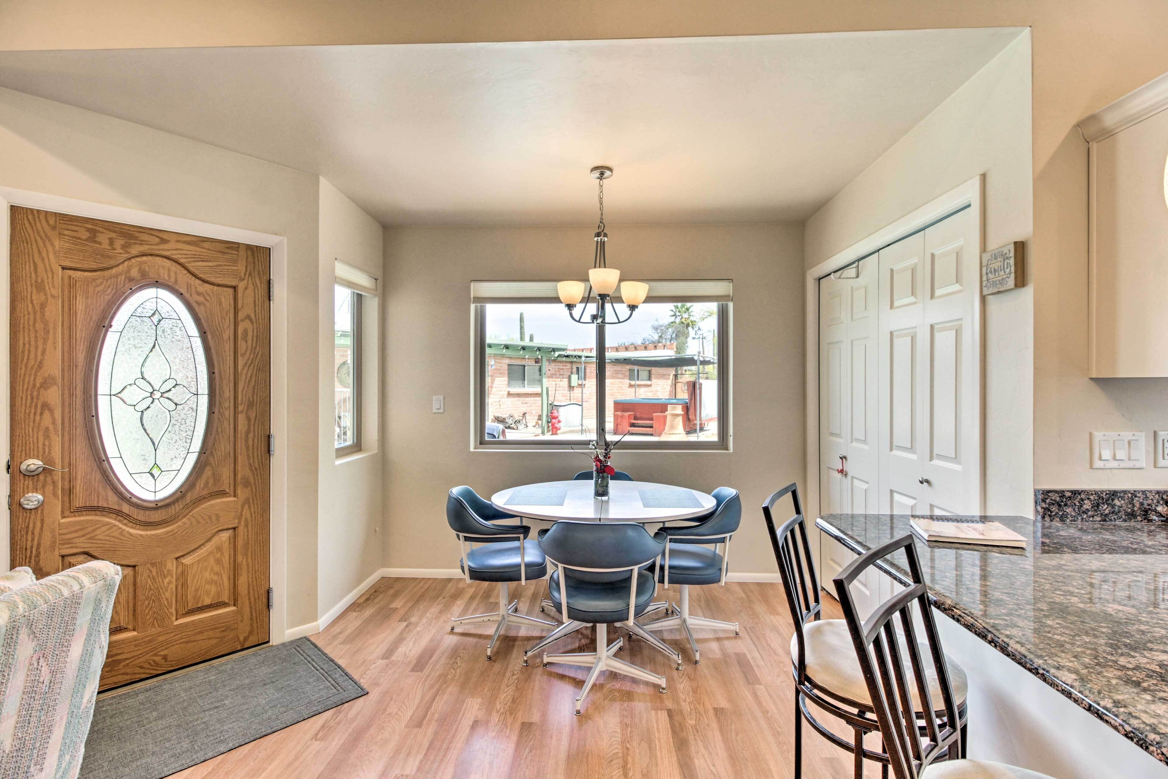 A dining area with a round table and six chairs, a wooden door with a stained glass window, and a view of the outside through a large window.