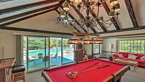 Une table de billard dans une pièce avec un canapé rouge, un lustre et une vue sur une piscine.