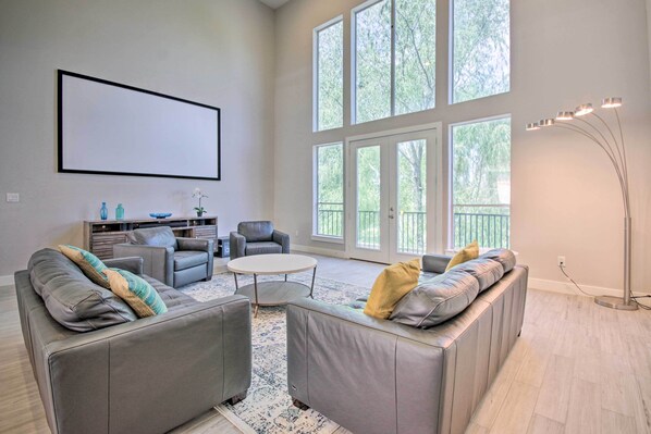 A spacious living room with a grey sectional sofa, a round coffee table, and a grey armchair. There is a large window providing natural light and a view of trees.