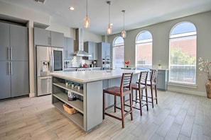A modern kitchen with grey cabinets, a stainless steel refrigerator, a large island with bar stools, and large windows with blinds.