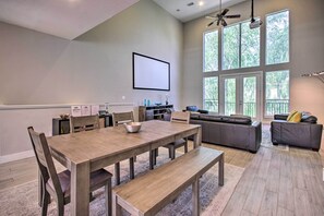 A spacious living room with a large dining table, comfortable seating, and a balcony with a view of trees.