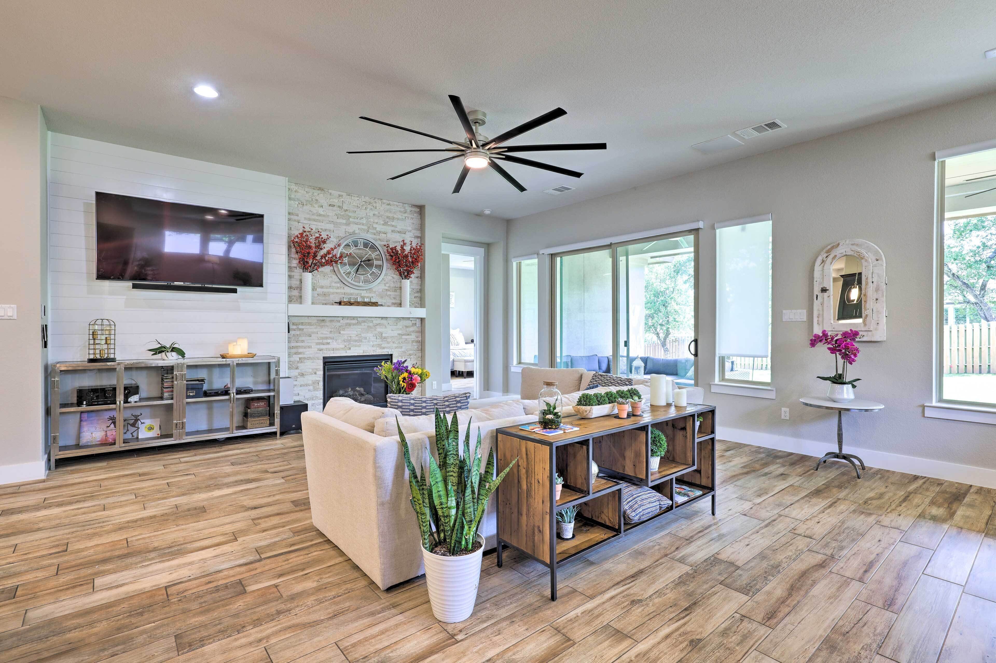 A spacious living room with a large flat-screen TV, a fireplace, a ceiling fan, and a large window providing natural light.