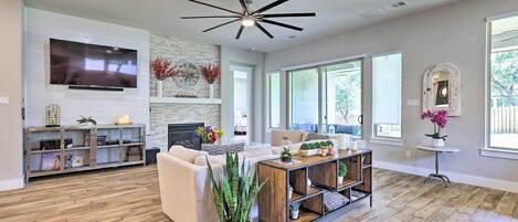 A spacious living room with a large flat-screen TV, a fireplace, a ceiling fan, and a large window providing natural light.