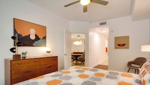 A bedroom with a bed, a dresser, a ceiling fan, a painting, and a dining area in the background. - Mid-century Modern Phoenix Condo, Walk to High St! (Phoenix)