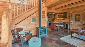 A wooden cabin interior with a staircase, leather furniture, a stone fireplace, and a kitchen area.