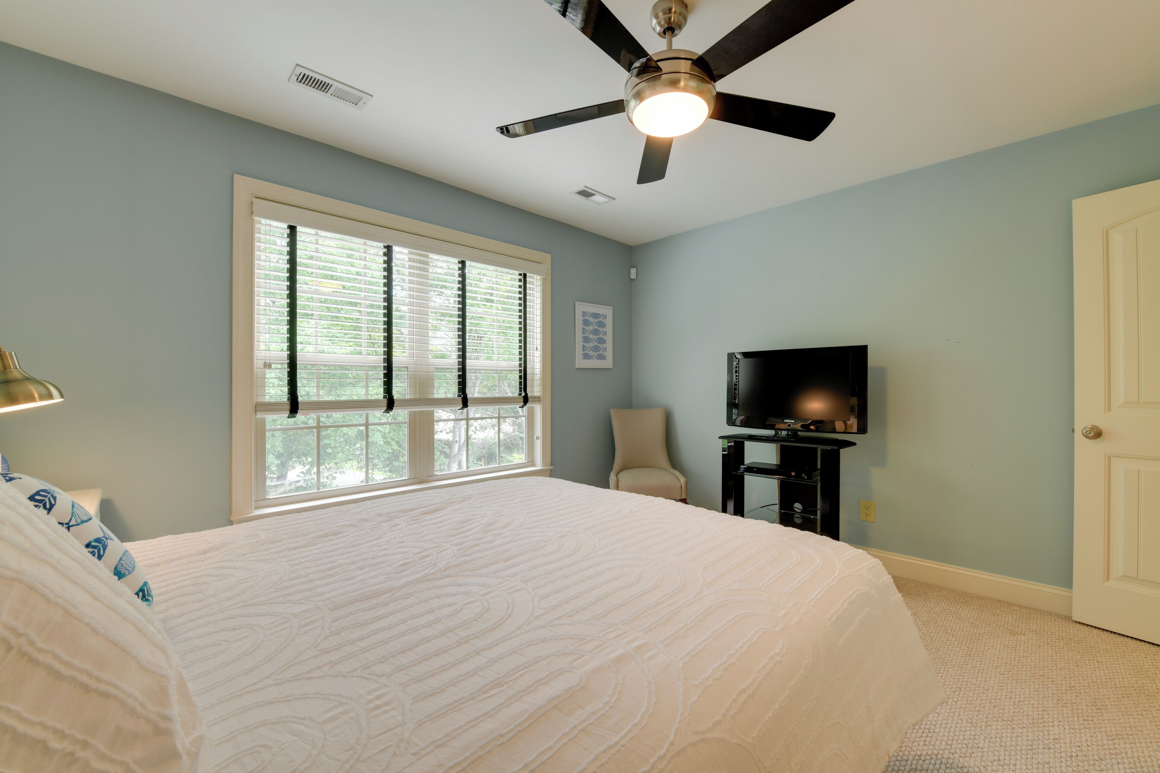 A bedroom with a large bed, a ceiling fan, a television, and a window with blinds.