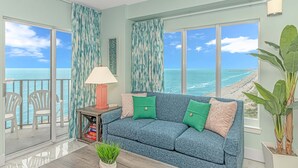 Sala de estar con un sofá azul, cojines verdes y beige, una mesa con una lámpara y vista a la playa.