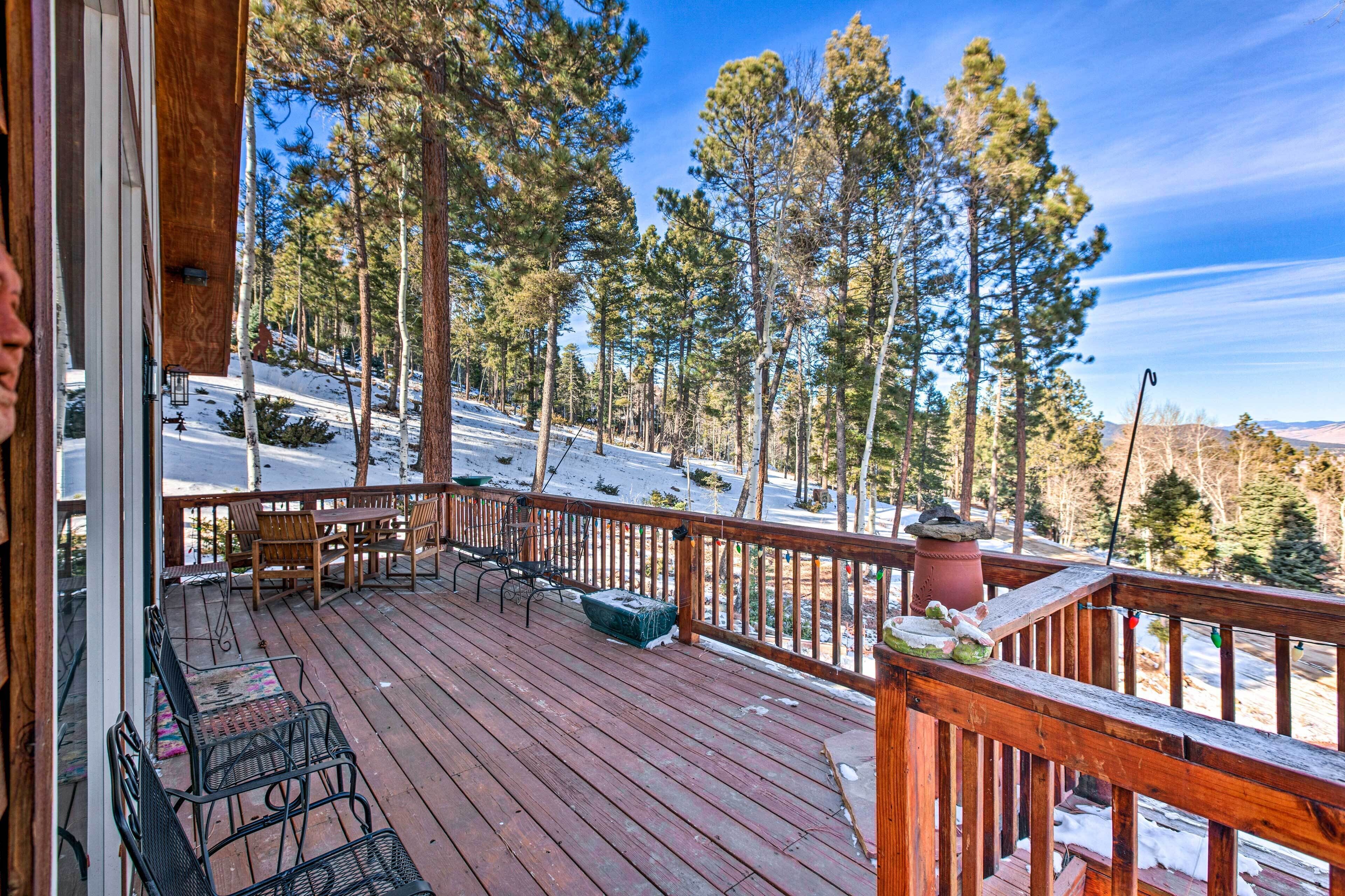 A wooden deck with outdoor seating, overlooking a snowy landscape and tall pine trees.
