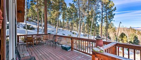 A wooden deck with outdoor seating, overlooking a snowy landscape and tall pine trees.