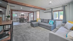 A modern living room with a grey sectional sofa, a small table, and a TV stand. There is a kitchen area in the background with white cabinets and a dining table.