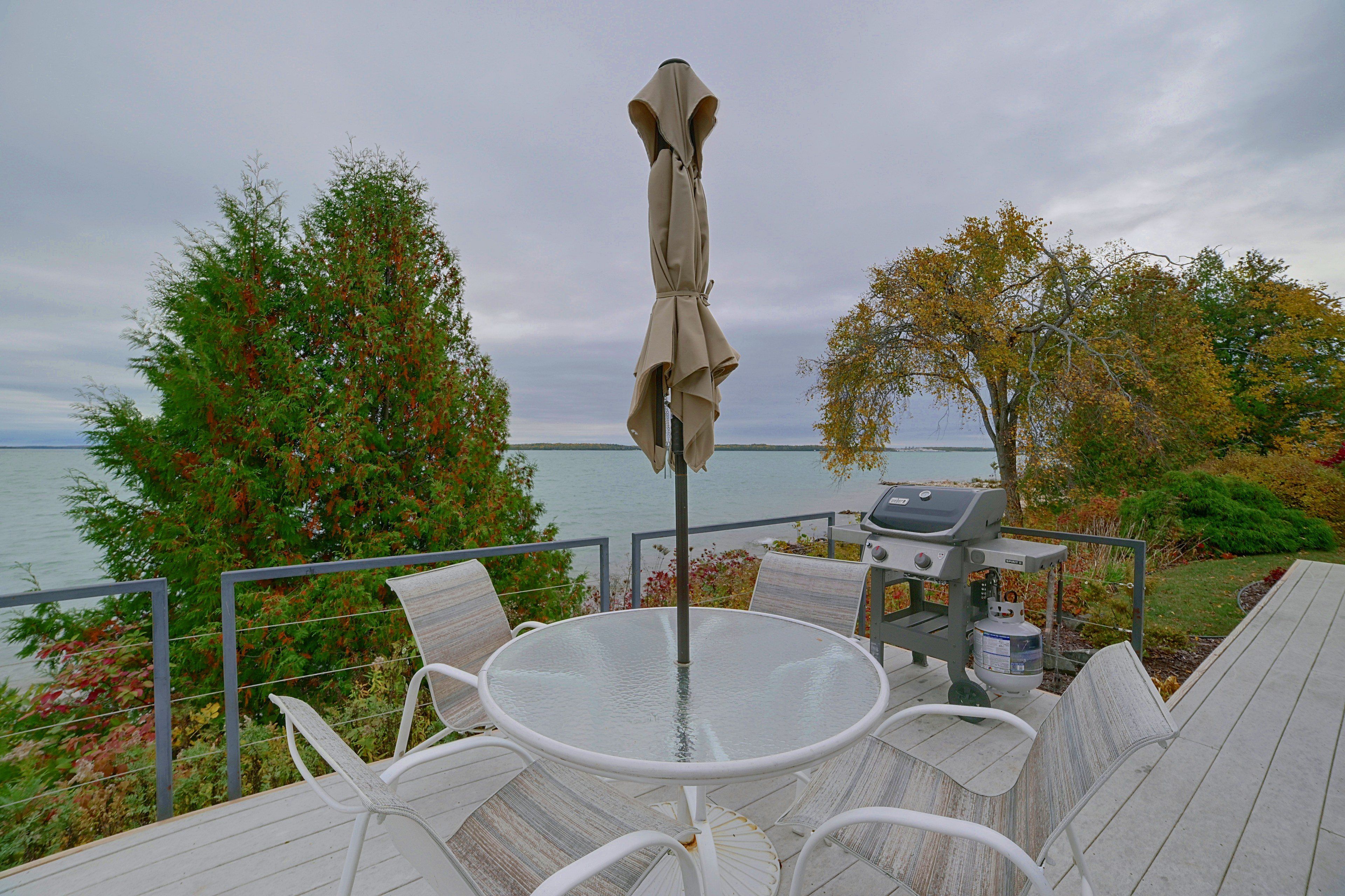 A deck with a round glass table and white chairs, overlooking a body of water with trees in the background.