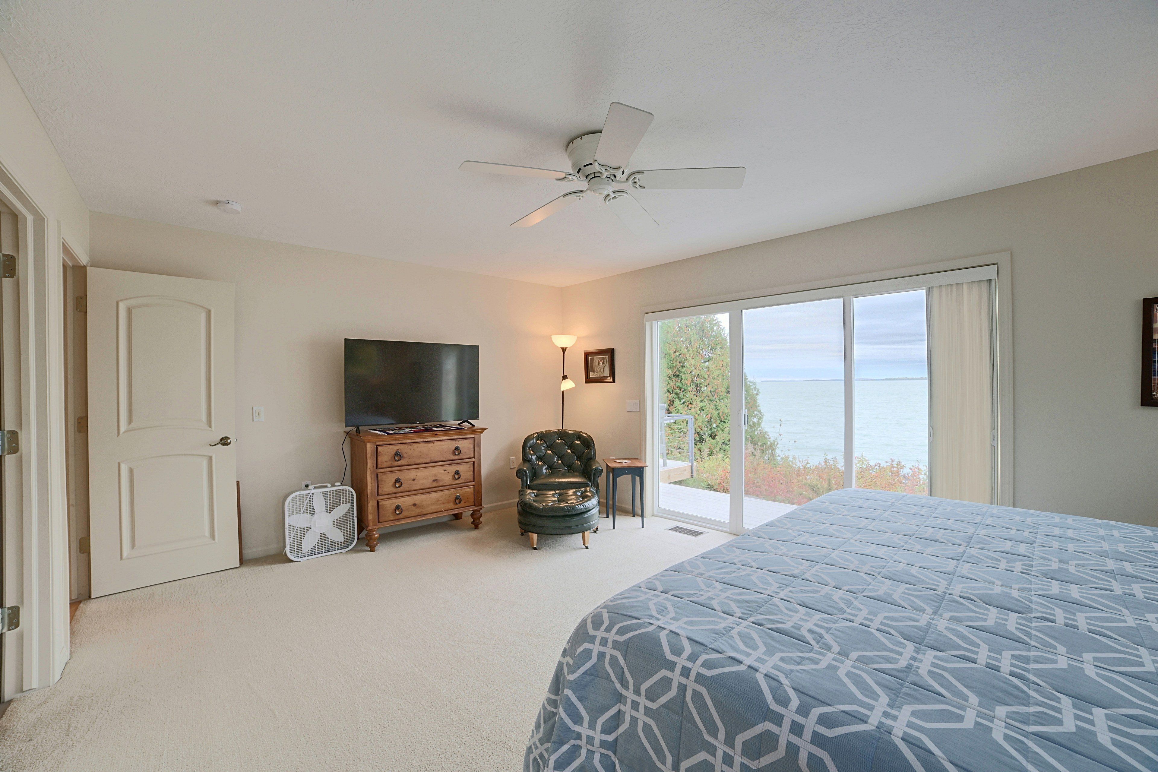 A bedroom with a bed, a dresser, a television, a chair, a fan, and a view of the outdoors.