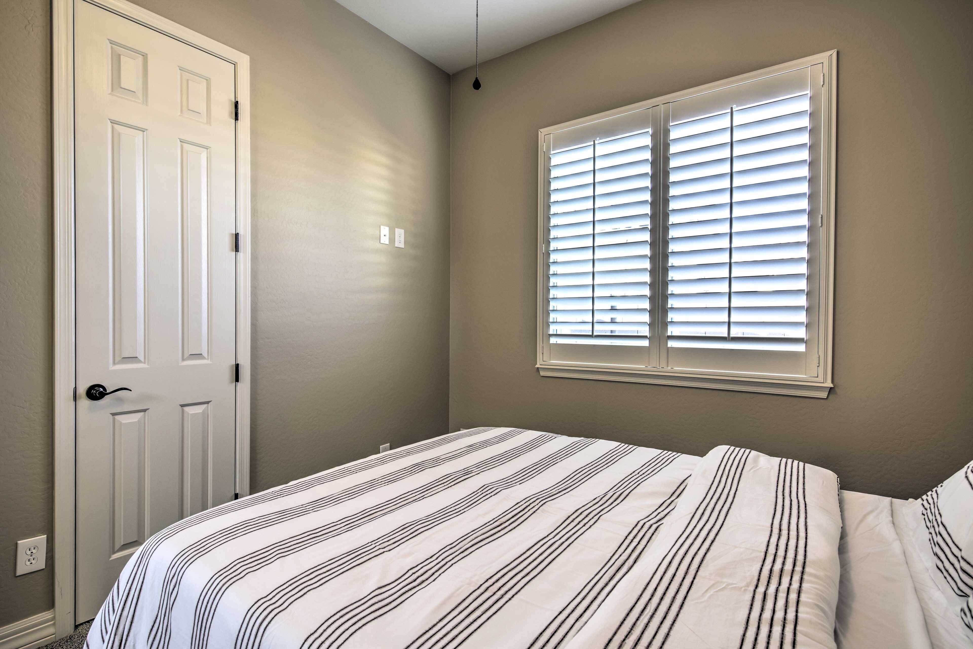 A bedroom with a bed, a window with blinds, a door, and a light switch on the wall.