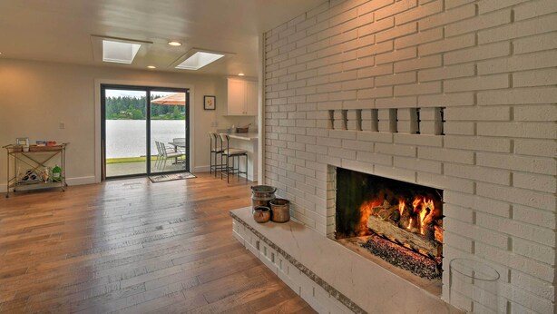 Amplia sala de estar con chimenea de ladrillo blanco, piso de madera y un gran ventanal que da a un área exterior.