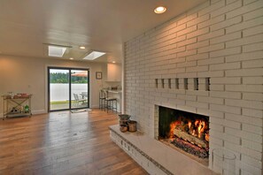 Ein geräumiges Wohnzimmer mit Kamin aus weißem Backstein, Holzboden und einer großen Glasschiebetür zum Außenbereich.