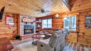 A cozy log cabin interior with a fireplace, leather sofa, and a ceiling fan. - Furnished Deck: Cabin in Coconino National Forest (Munds Park)