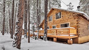A log cabin with a wooden deck and a chimney, surrounded by snow-covered trees. - Furnished Deck: Cabin in Coconino National Forest (Munds Park)