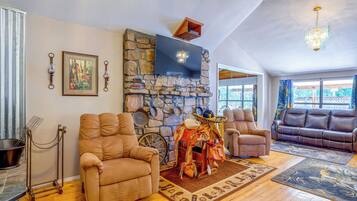 A spacious living room with a stone fireplace, multiple seating options including a recliner and a sofa, and a large flat-screen TV mounted above the fireplace.