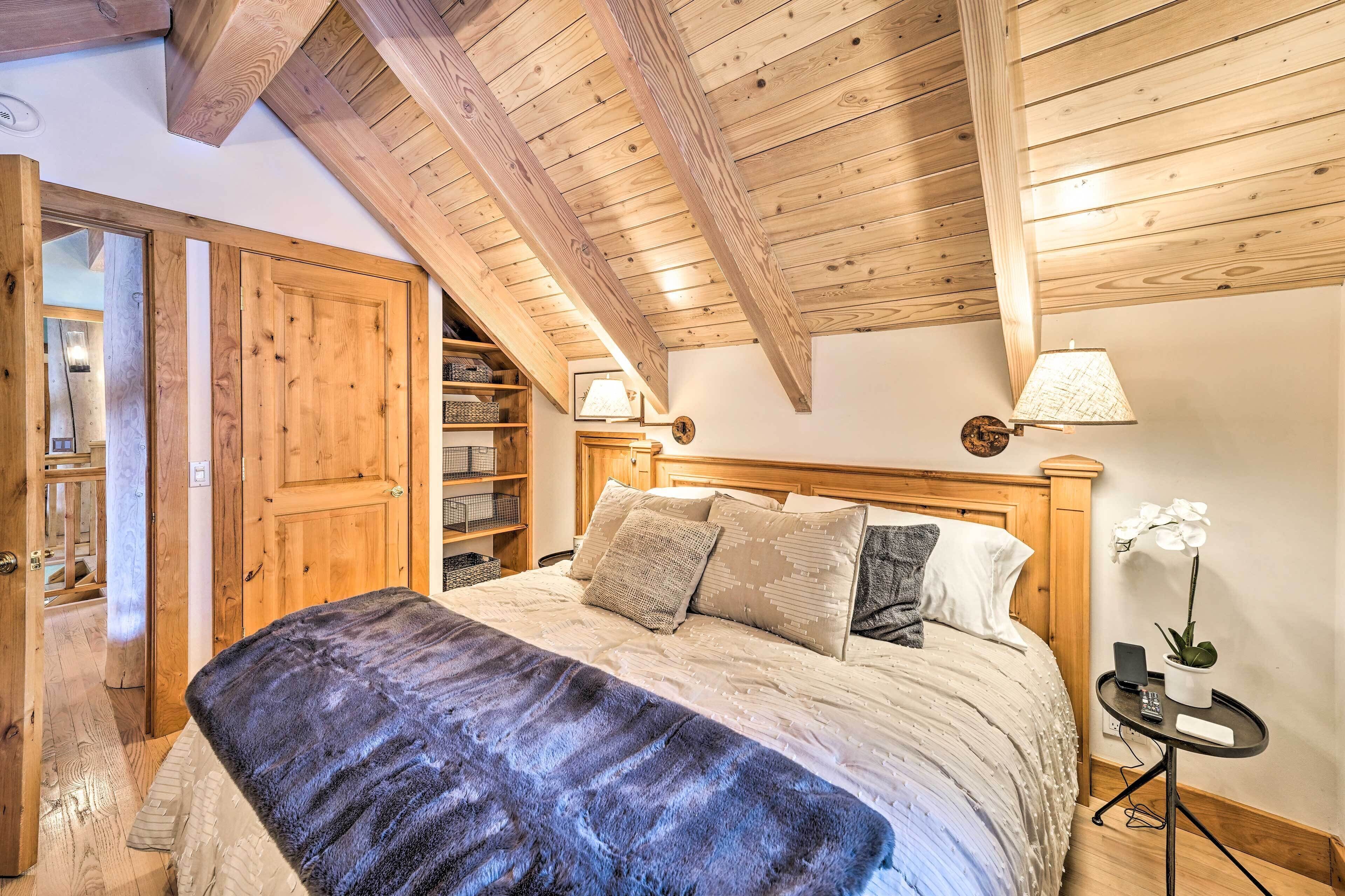 A bedroom with a wooden ceiling, a bed with a blue blanket, a bedside table with a lamp, and a small plant.
