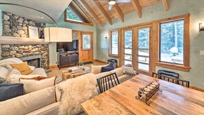 Un salon chaleureux avec une cheminée en pierre, des poutres en bois au plafond et de grandes fenêtres donnant sur des arbres couverts de neige.