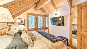 Une chambre avec un plafond en pente, des poutres en bois, un lit recouvert de plusieurs coussins, un banc et une porte donnant sur une autre pièce.