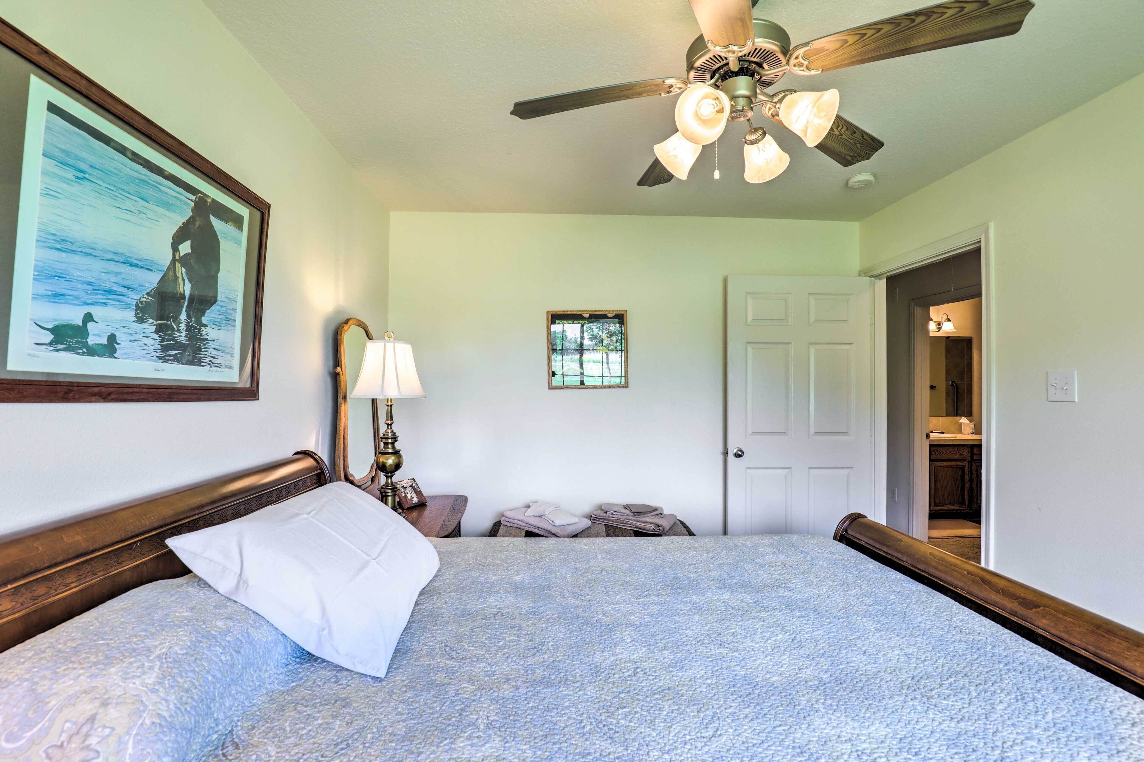 Une chambre à coucher comprenant un lit, un ventilateur de plafond, un tableau encadré, une lampe et une porte donnant sur une salle de bain.