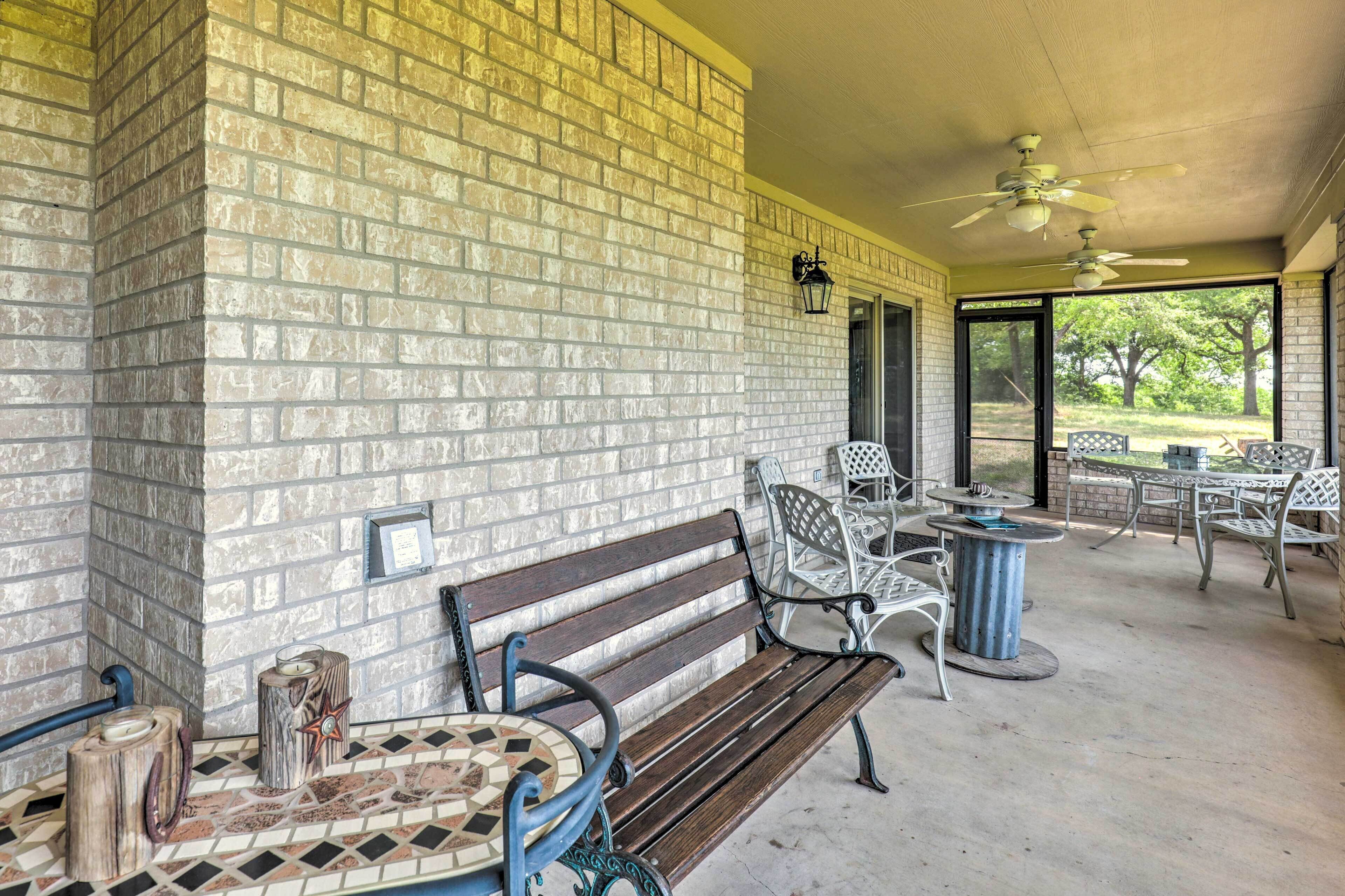 Een overdekt terras met een bakstenen muur, een houten bank, metalen stoelen, een tafel en plafondventilatoren.
