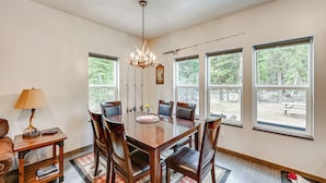Comedor con mesa de madera, seis sillas, una lámpara de araña y ventanas amplias con vista a árboles.