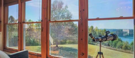 A room with large windows, a telescope, and a view of a lake and trees.