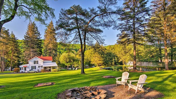 Une maison avec un toit rouge, un foyer extérieur et deux chaises Adirondack dans une cour verdoyante, entourée d’arbres.