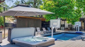 A hot tub on a wooden deck with a pool, lounge chairs, and a basketball hoop.