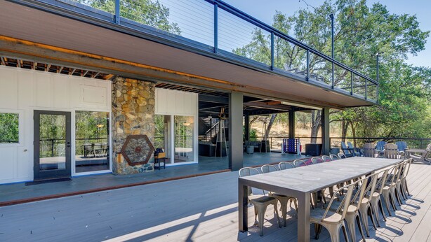 An outdoor deck with a long table and chairs, a stone wall, and a covered area with a railing.