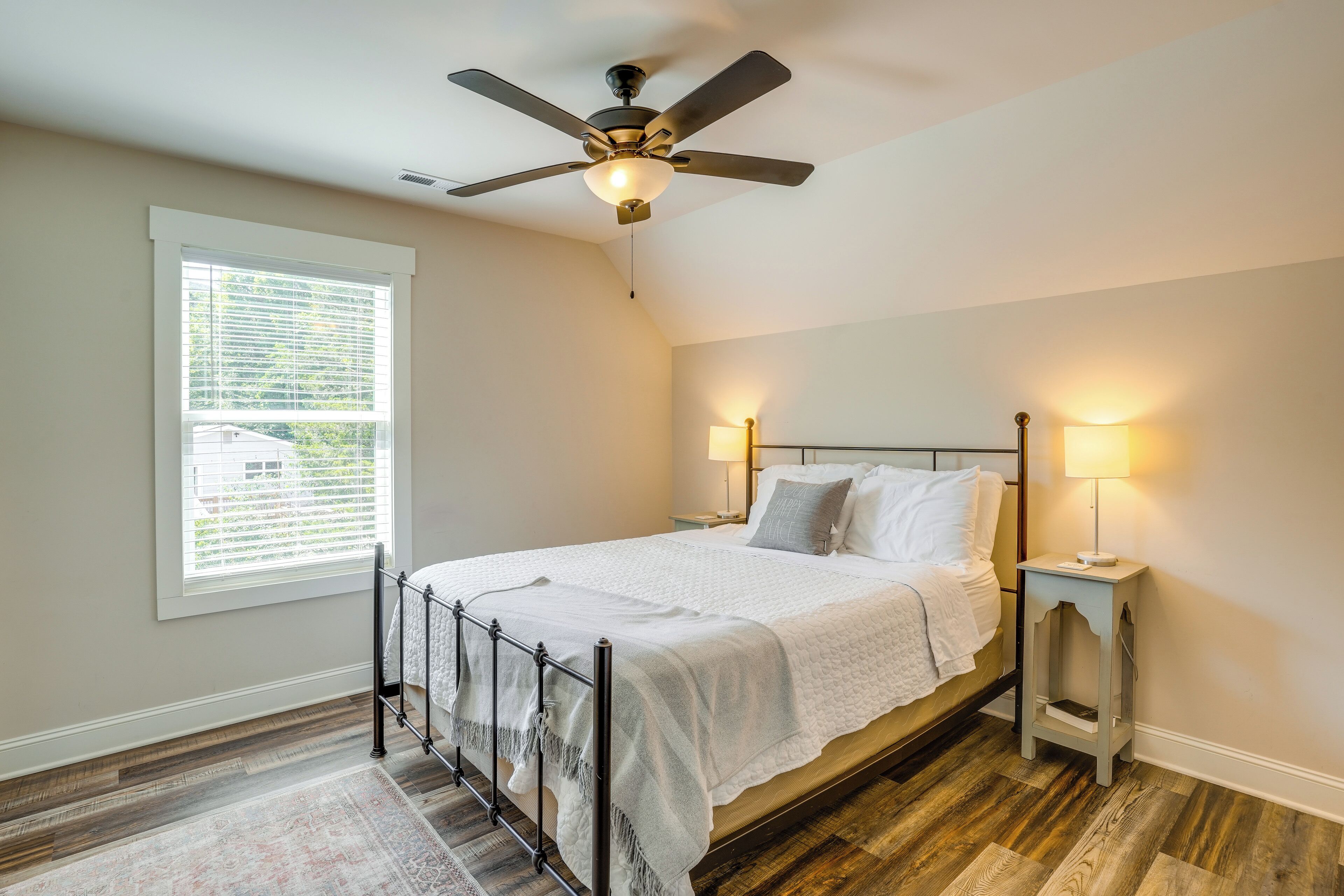 A bedroom with a bed, a ceiling fan, bedside lamps, a window, and a wooden floor.
