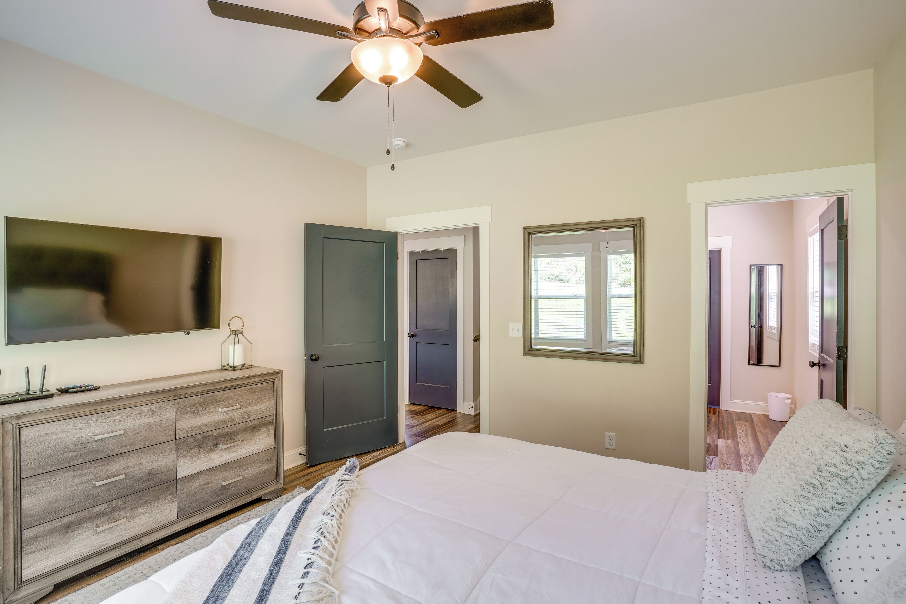 A bedroom with a bed, a dresser, a ceiling fan, a TV, and a door leading to another room.