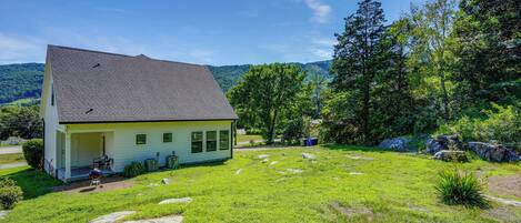 A house with a sloped roof, a porch, and a grassy yard with rocks and trees.