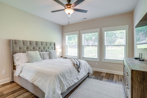 A bedroom with a tufted headboard, a bed with white bedding, a ceiling fan, and three windows with blinds. - Chattanooga Mtn Retreat, Walk to The Venue! (Chattanooga)