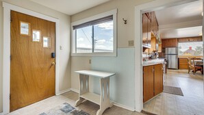 A hallway with a wooden door, a small table, a window with blinds, and a kitchen area visible through an open door. - Charming Trinidad Home ~ 2 Mi to Main Street! (Trinidad)