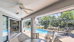 A sunlit patio with a glass table and chairs, a ceiling fan, and a view of a swimming pool and outdoor seating area. - Modern House < 5 Miles to Port Charlotte Beach! (Port Charlotte)