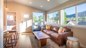 A modern living room with a brown leather sofa, a wooden coffee table, and a blue armchair. There are large windows providing a view of the outdoors. - Golden Cottage Near Red Rocks & School of Mines! (Golden)