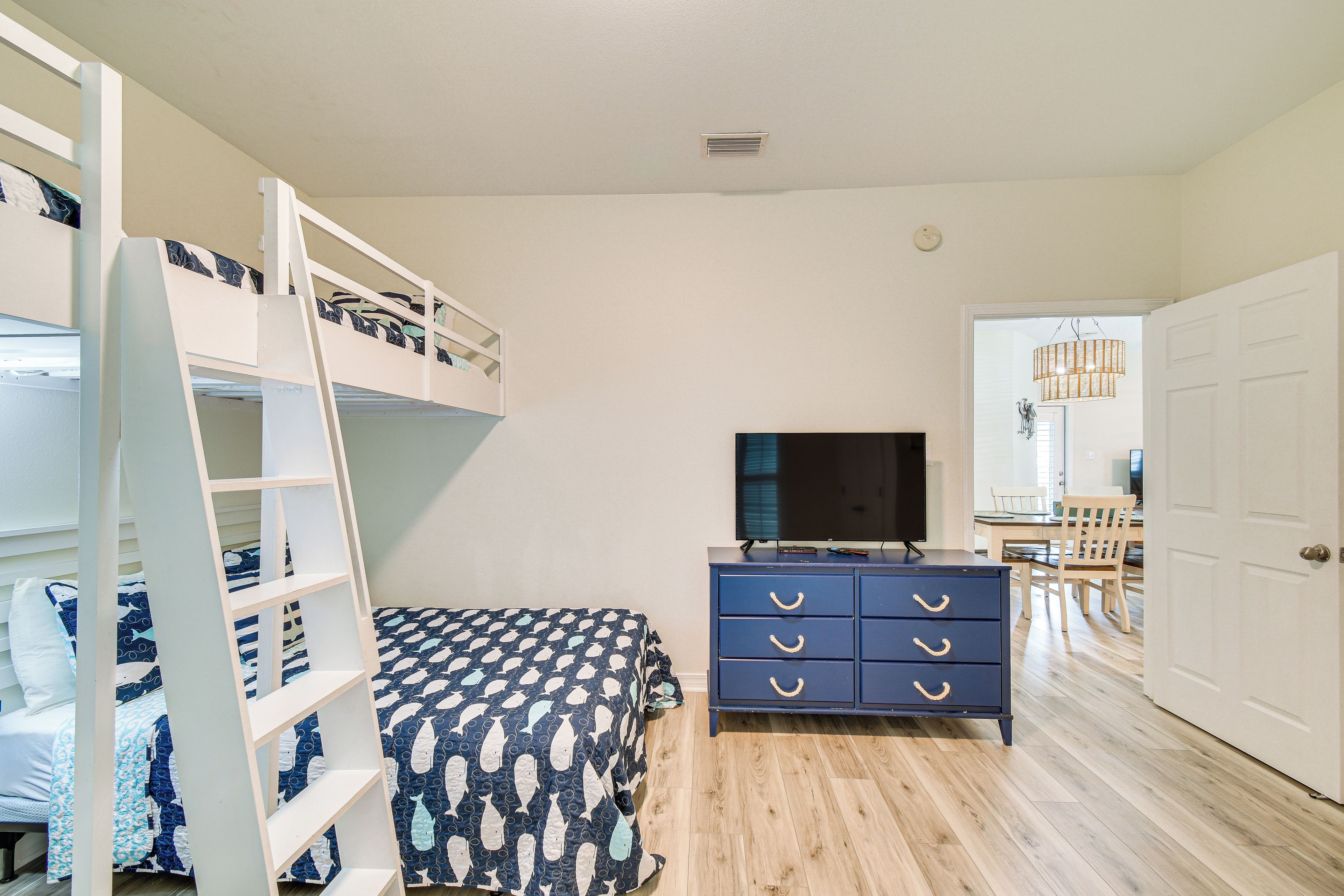A bedroom with bunk beds, a TV, a dresser, and a dining area visible through an open door.