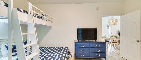 A bedroom with bunk beds, a TV, a dresser, and a dining area visible through an open door.