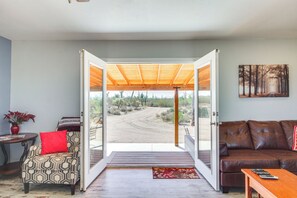 An open door leading to a covered outdoor area with a dirt path, a brown leather sofa, a patterned armchair with a red pillow, and a small table with a vase of flowers. - Secluded Marana Home w/ Viewing Decks + Privacy! (Marana)