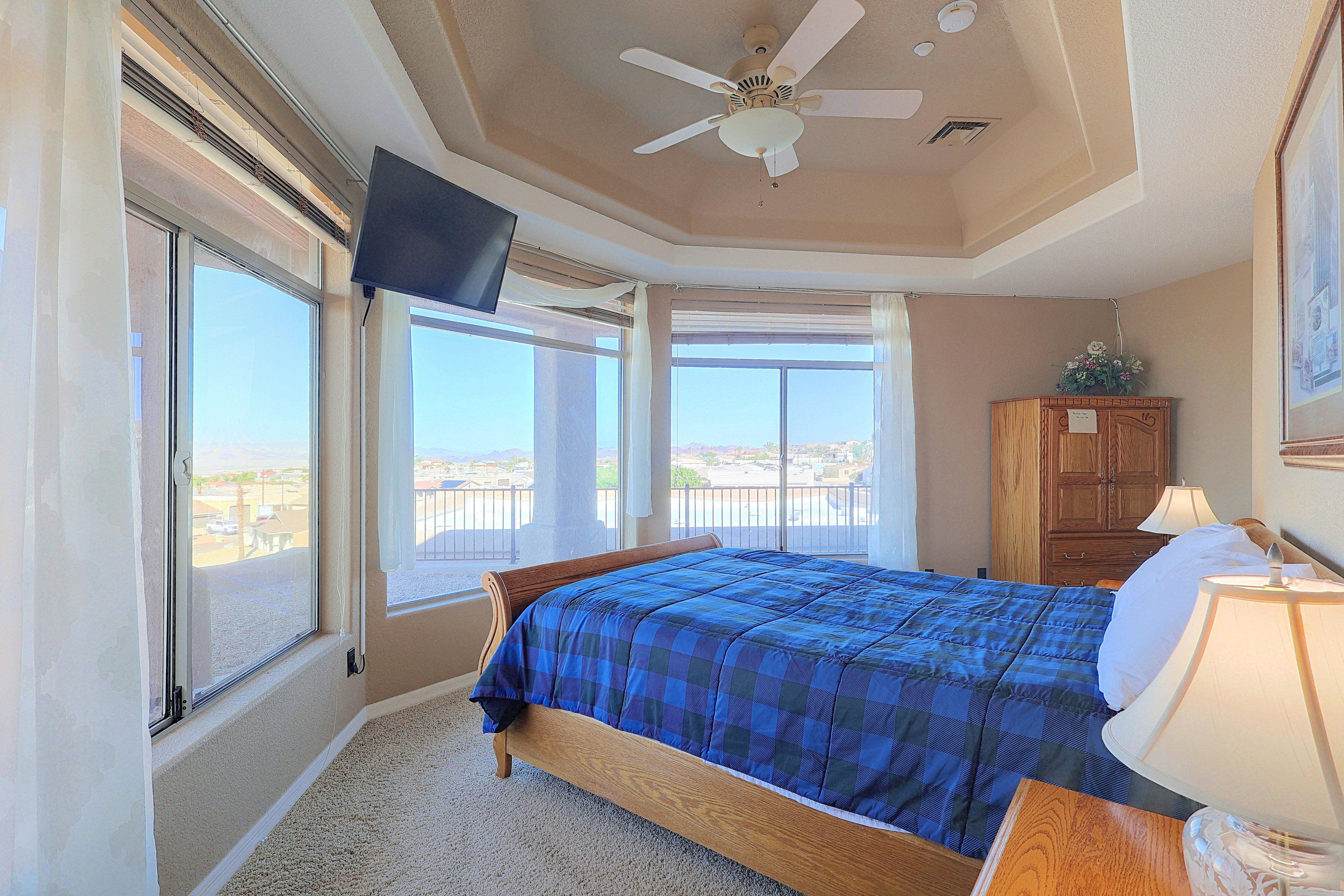 A bedroom with a bed, a TV mounted on the wall, a ceiling fan, and a view of the outside through large windows.