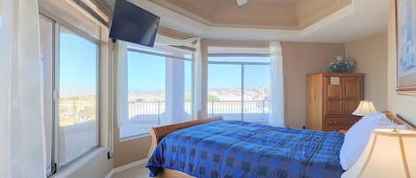 A bedroom with a bed, a TV mounted on the wall, a ceiling fan, and a view of the outside through large windows.